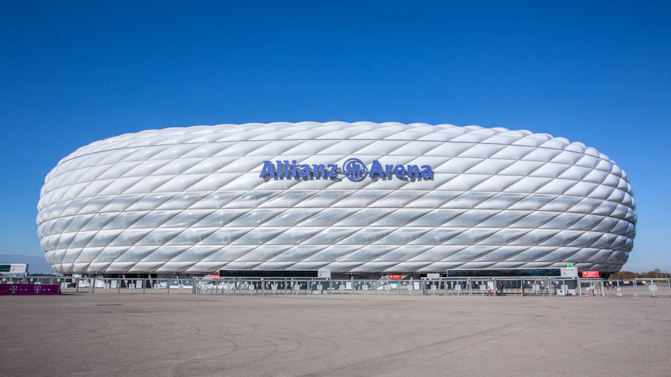 Large white stadium labeled "Allianz Arena" against a clear blue sky with an expansive empty pavement in the foreground.