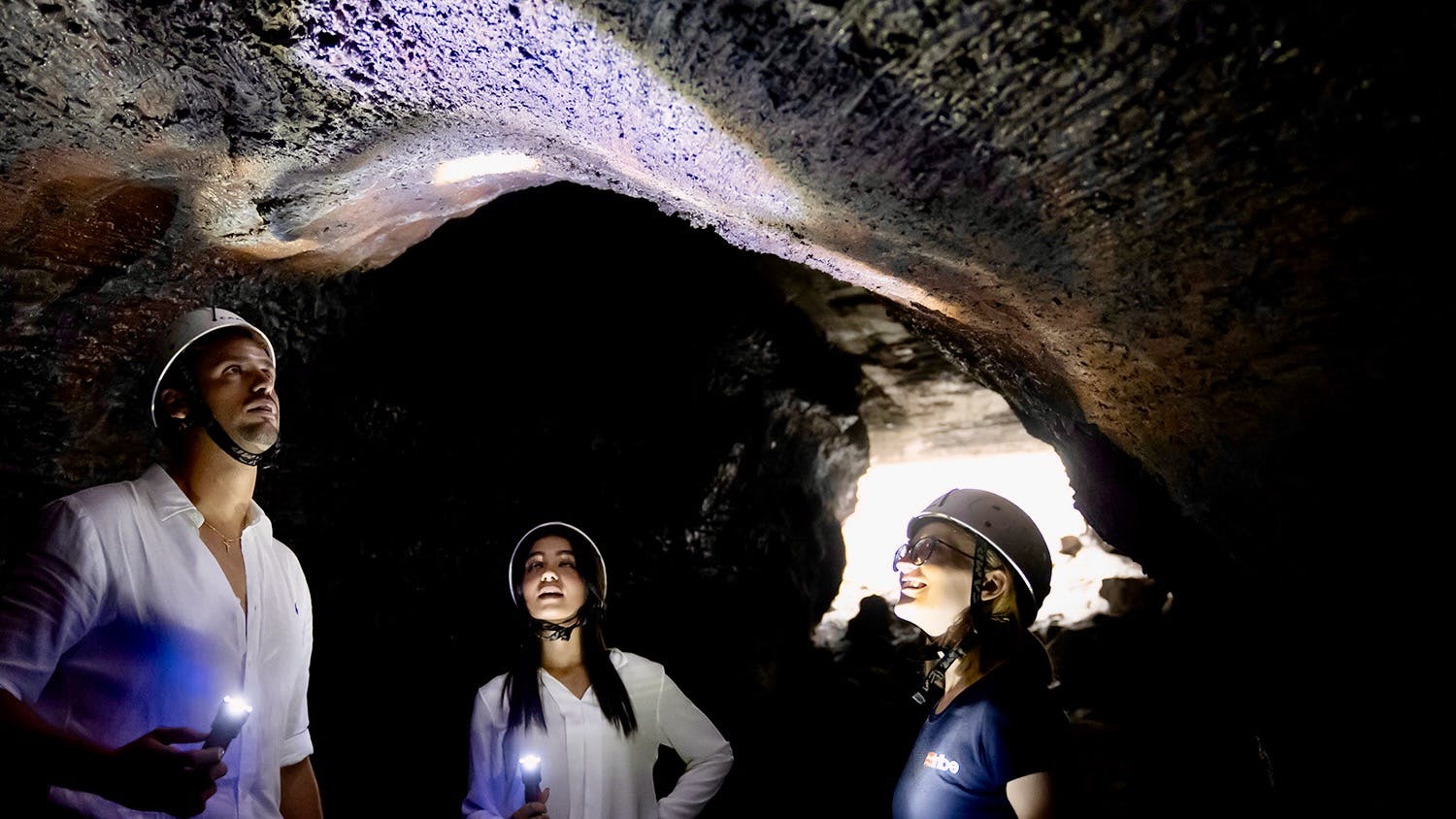Τρία άτομα που φορούν κράνη και προβολείς εξερευνούν μια σπηλιά με χαμηλό φωτισμό, χρησιμοποιώντας φακούς για να φωτίσουν την οροφή της σπηλιάς.