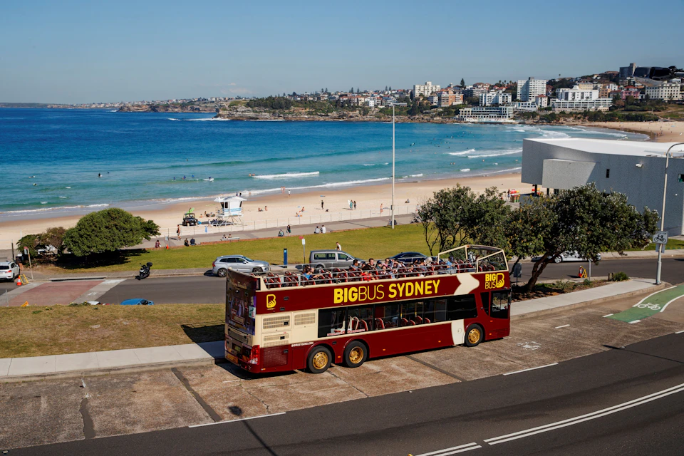 A red double-decker bus labeled "Big Bus Sydney" parked near a beach with people, ocean, and buildings in the background.