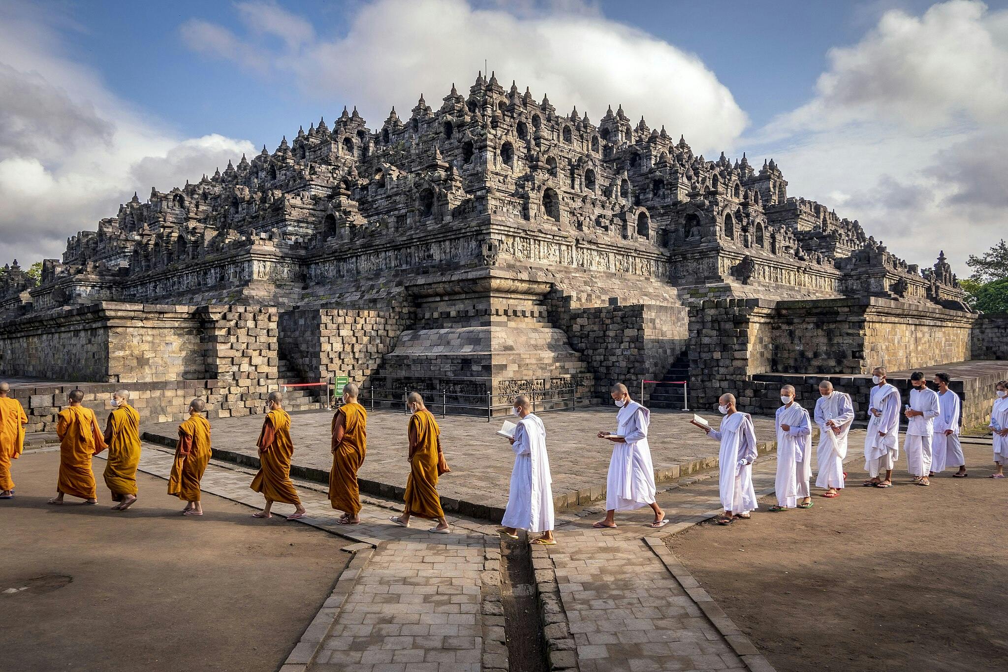 A line of monks and nuns in orange and white robes walk in front of the ancient Borobudur temple on a sunny day.