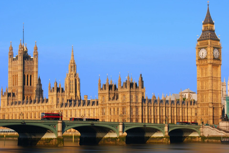 The Palace of Westminster with its gothic architecture, seen from across the Thames River under a clear blue sky.