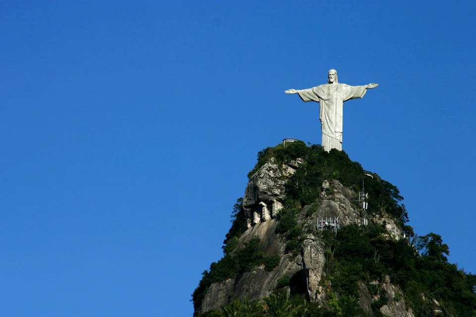 A large statue of Christ with outstretched arms stands atop a rocky, vegetation-covered hill against a clear blue sky.
