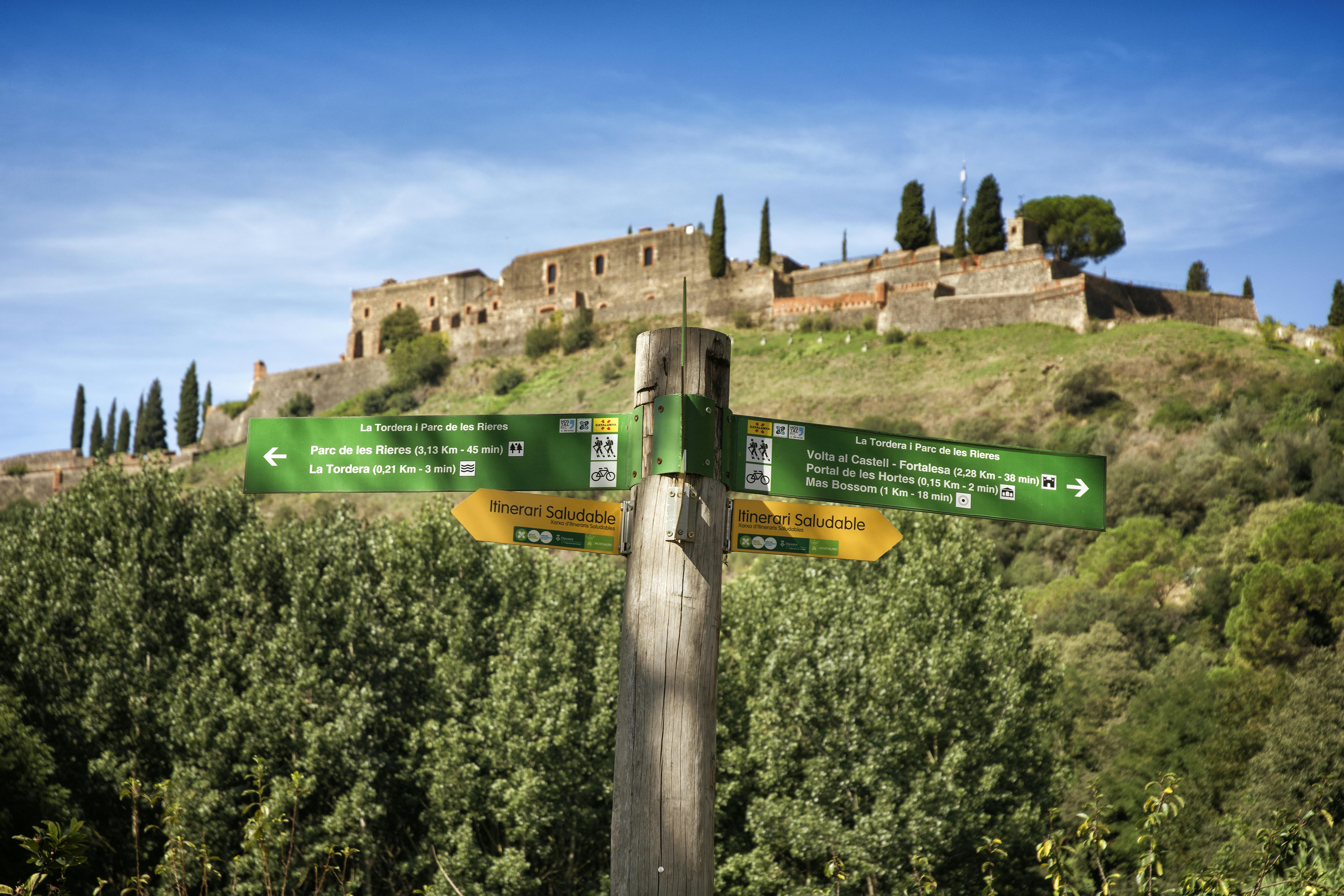 Wooden signpost with green and yellow directional signs in front of an old stone fort on a grassy hill under a blue sky.