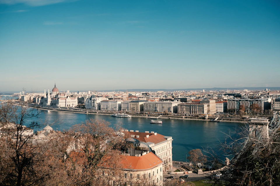 A cityscape featuring a wide river, historic buildings, and a prominent dome-shaped structure under a clear blue sky.