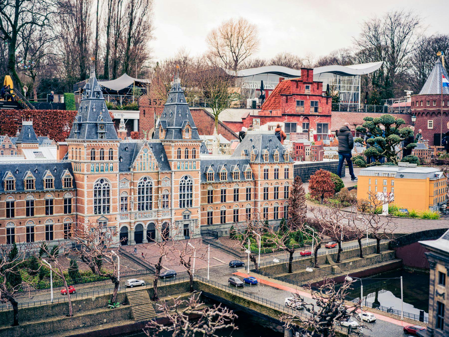 大きな華やかな建物や周囲の家など、細かいミニチュアの建物の隣に人が立っており、近くには裸の木や模型車があります。