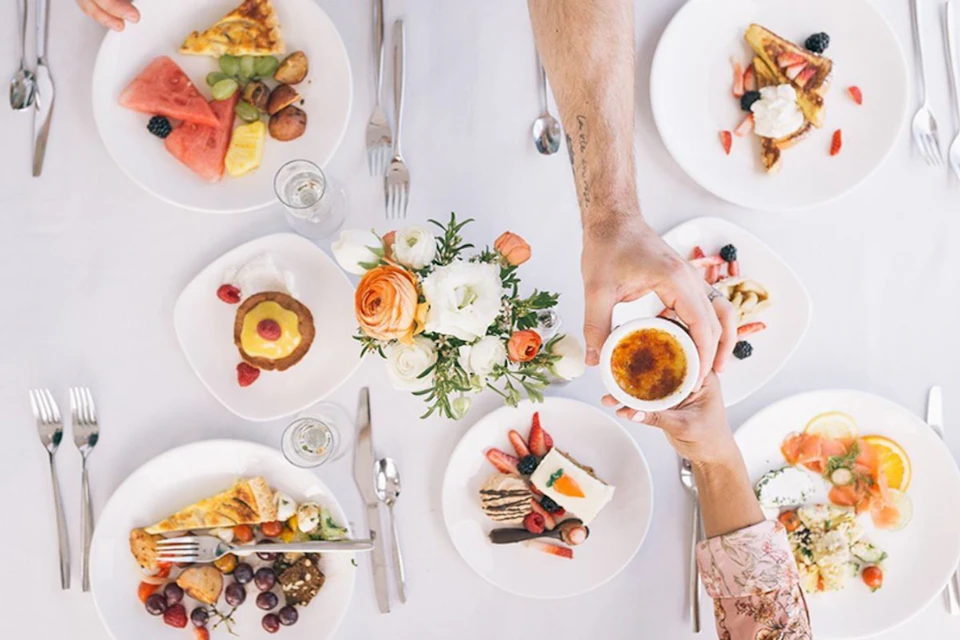 Aerial view of a table with plates of assorted desserts and fruits, a flower centerpiece, and hands reaching for a crème brûlée.