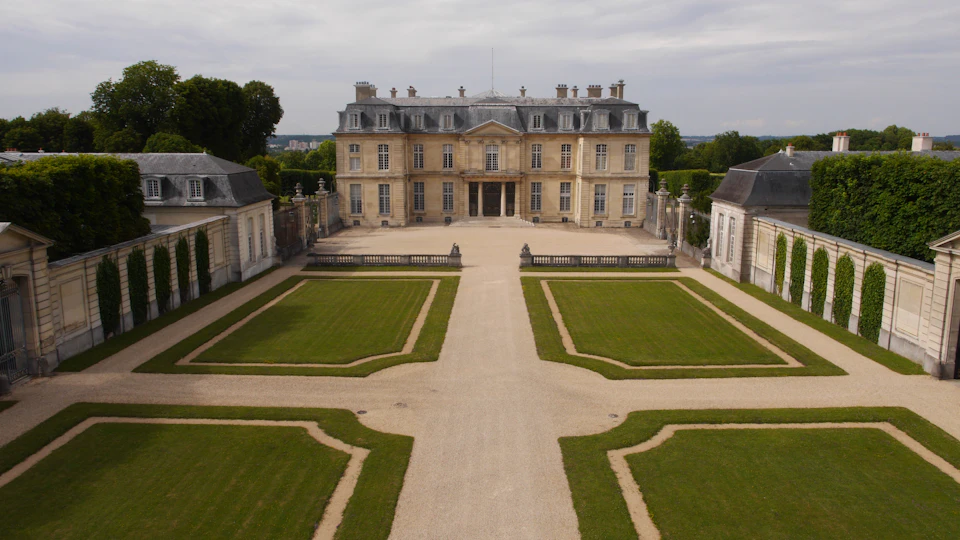 A large, stately mansion with a symmetrical design is surrounded by meticulously manicured lawns and pathways.