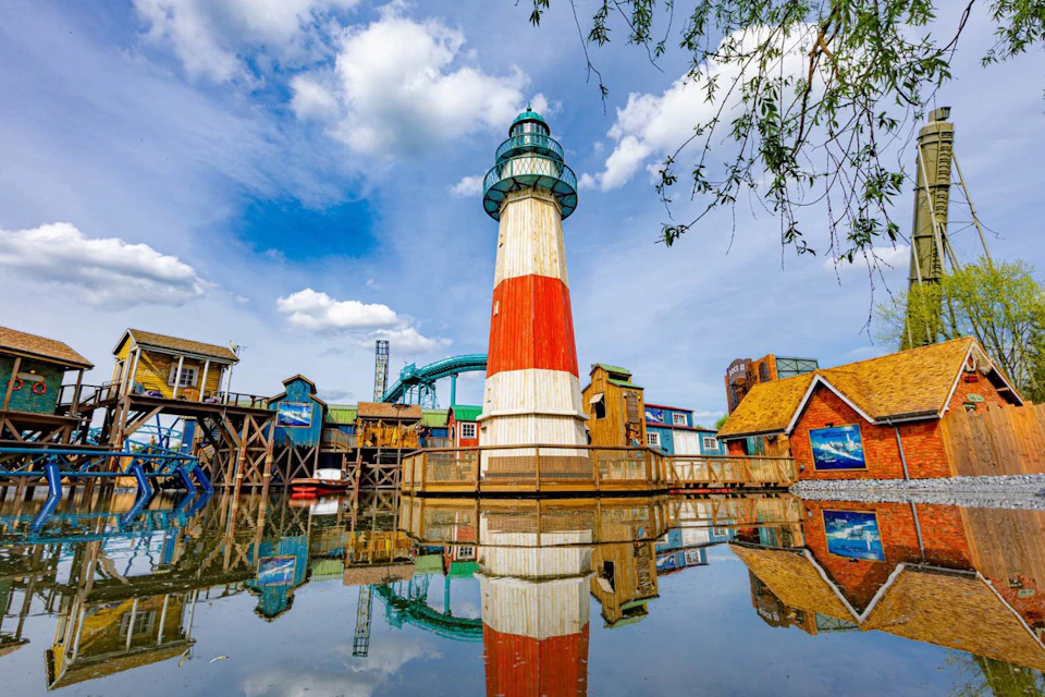 A colourful lighthouse and buildings are reflected in the still water beneath a partly cloudy sky, with greenery framing the scene.