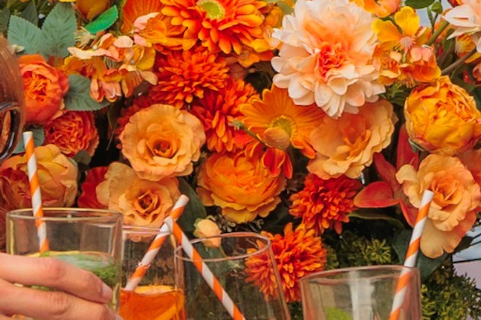 A group of people enjoying drinks on a boat decorated with orange flowers, set on a canal lined with trees and buildings.