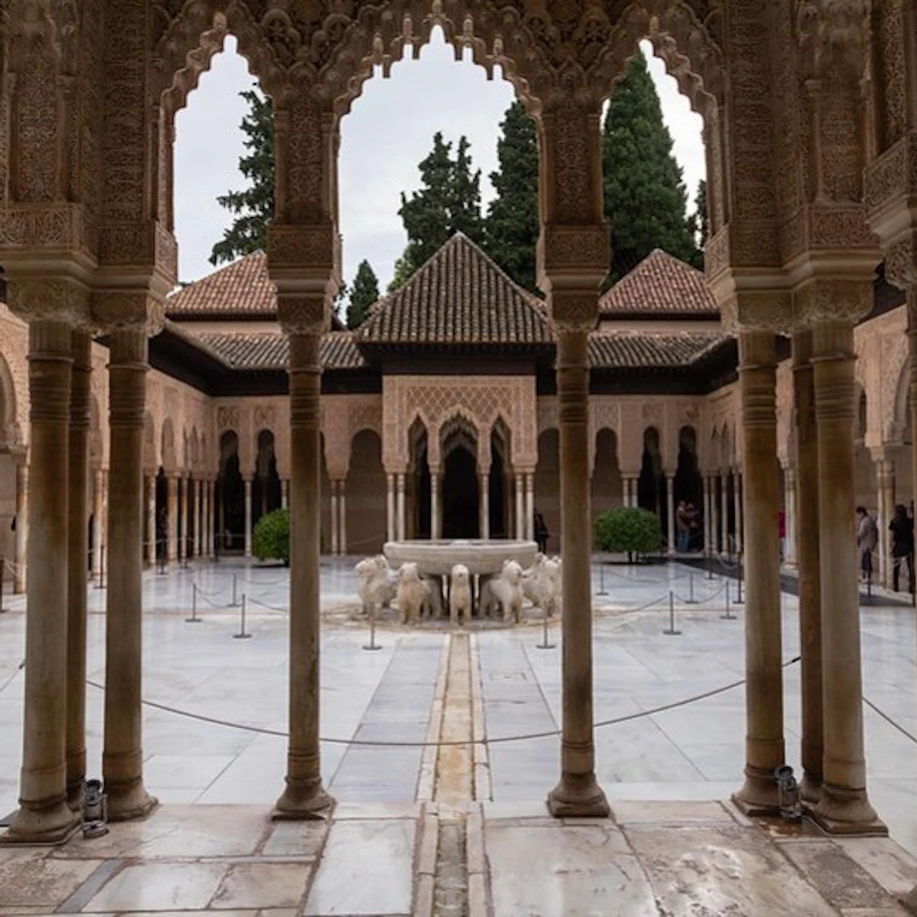 Ornate courtyard with arched columns, lion fountain centerpiece, intricate carvings, and surrounding tiled roofs. Trees in the background.