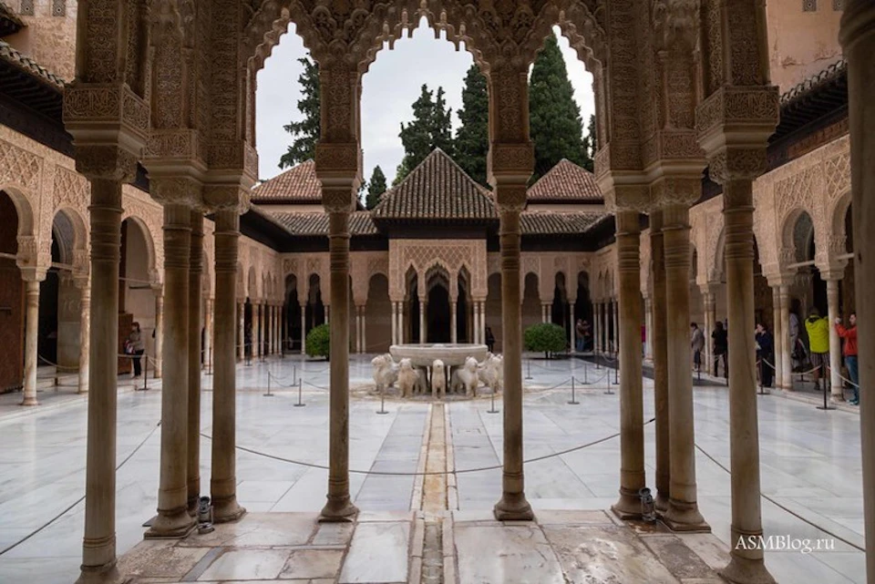 Ornate courtyard with arched columns, lion fountain centerpiece, intricate carvings, and surrounding tiled roofs. Trees in the background.