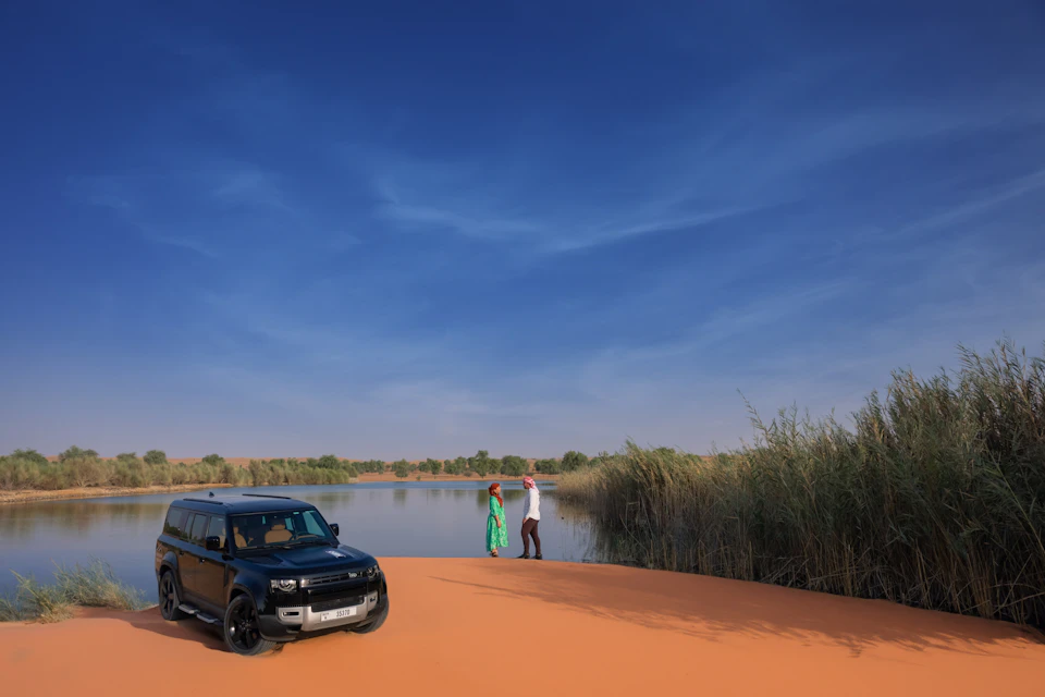 A black SUV is parked beside a small lake. Two people stand near the water on the sandy shore under a blue sky.
