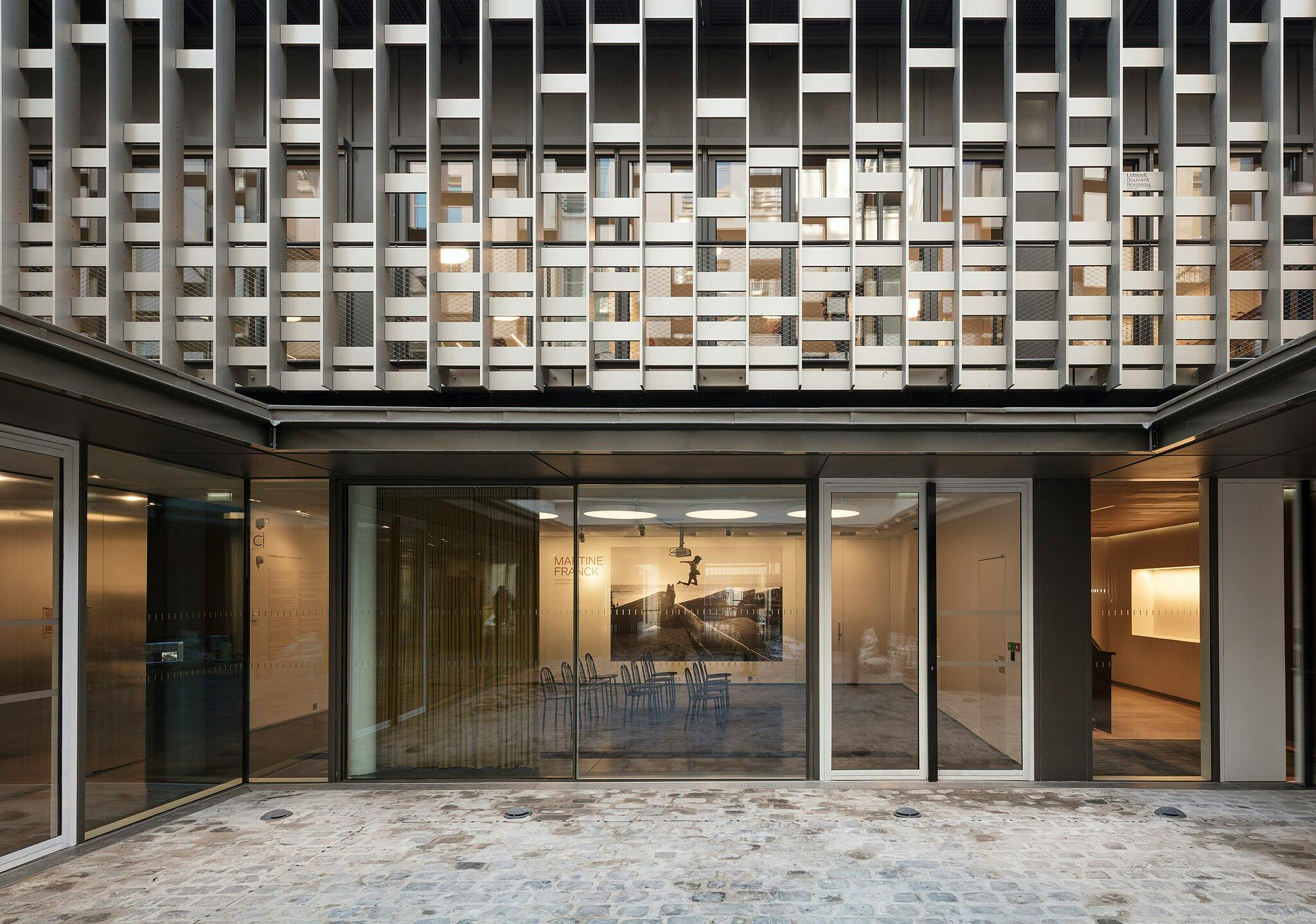 Modern building with large glass windows, showing a small interior room with chairs, light fixtures, and a black-and-white photograph.