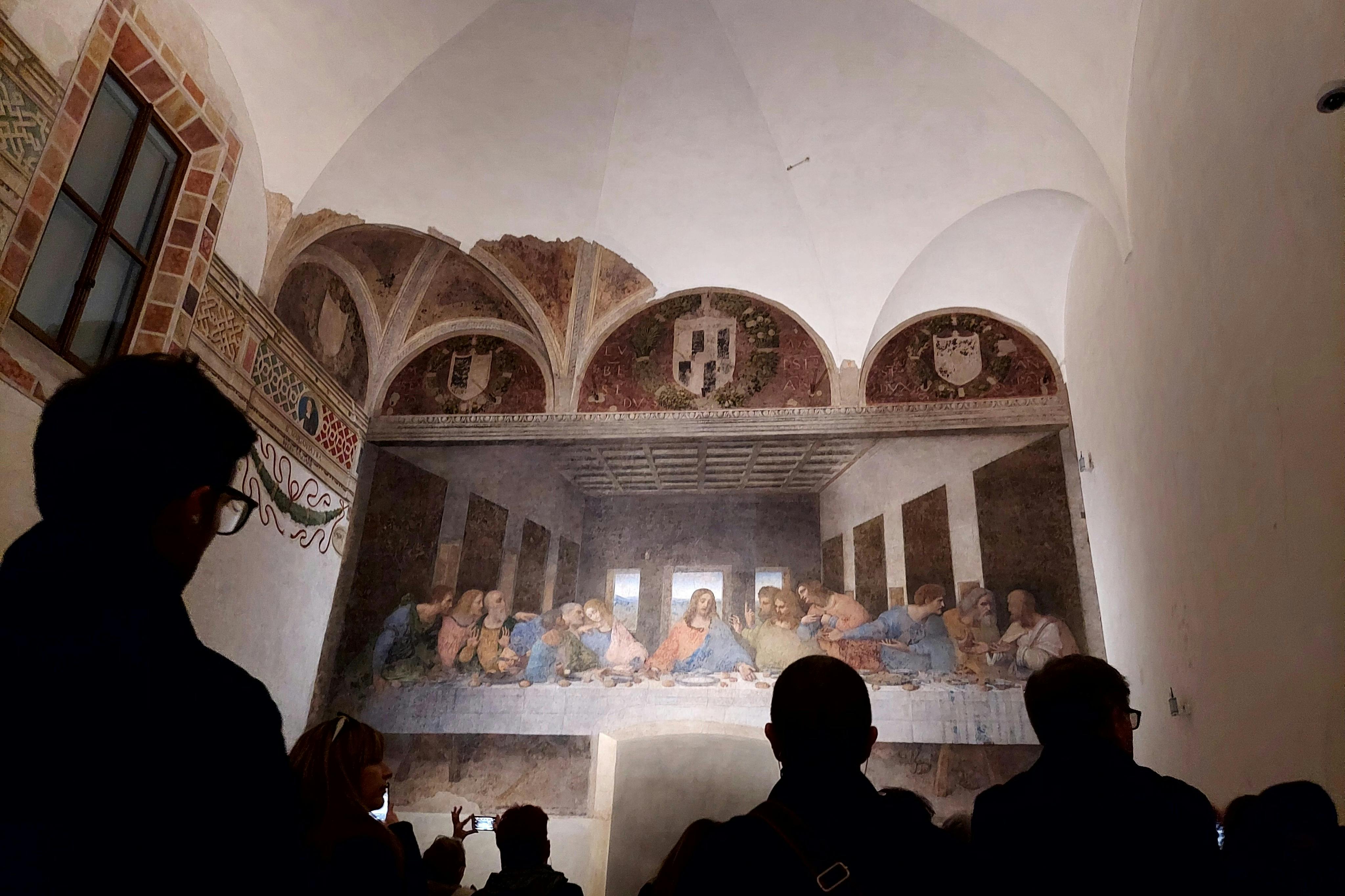 A group of people view a large fresco painting of The Last Supper on a wall in an arched room.