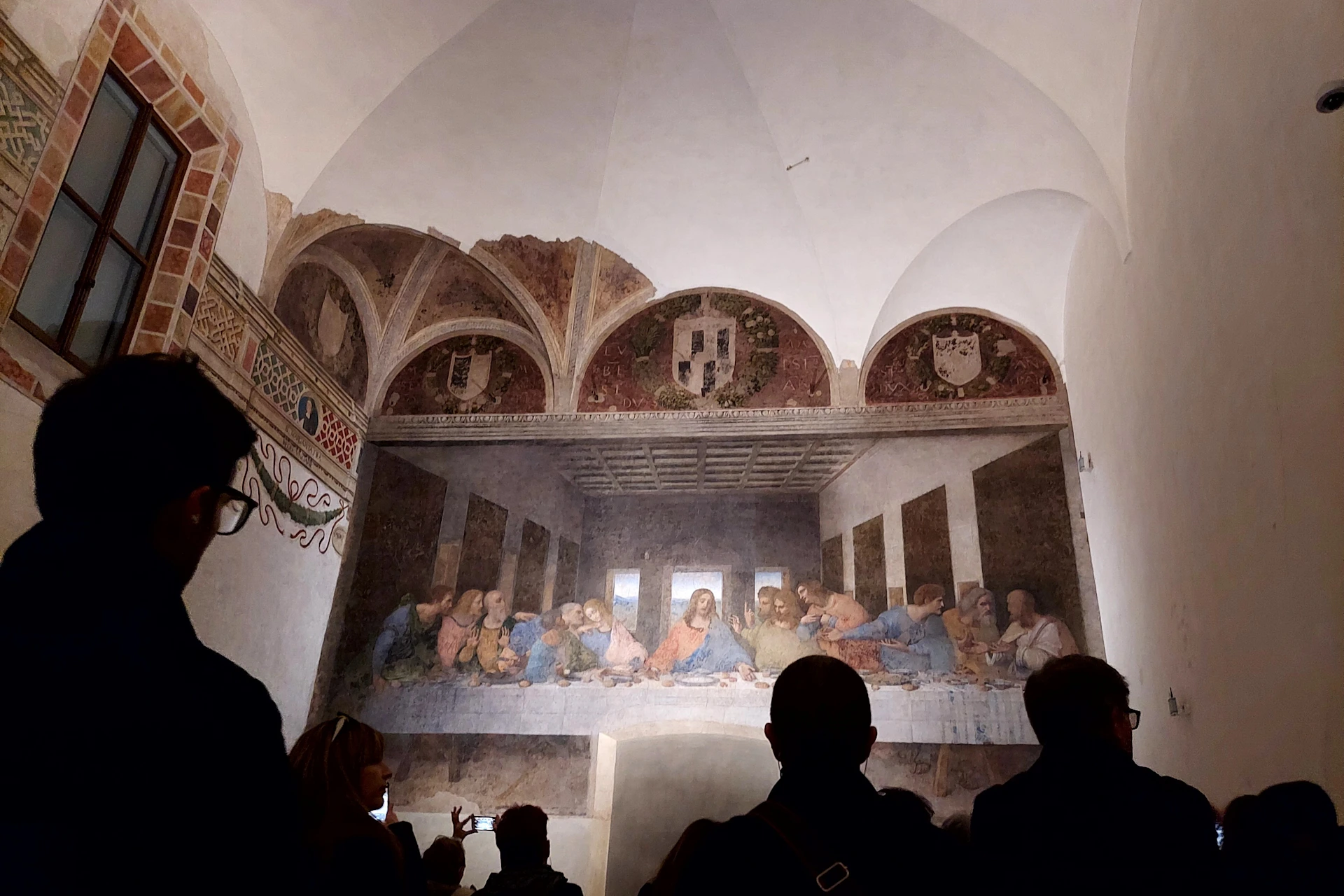 A group of people view a large fresco painting of The Last Supper on a wall in an arched room.