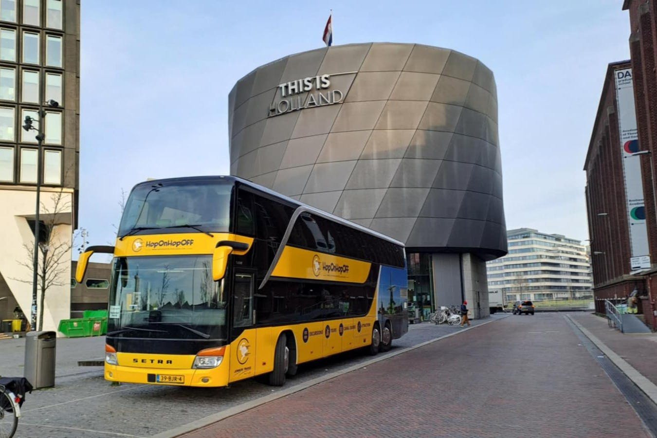 A yellow double-decker bus labeled "Hop On Hop Off" is parked in front of a modern building with "This is Holland" signage.