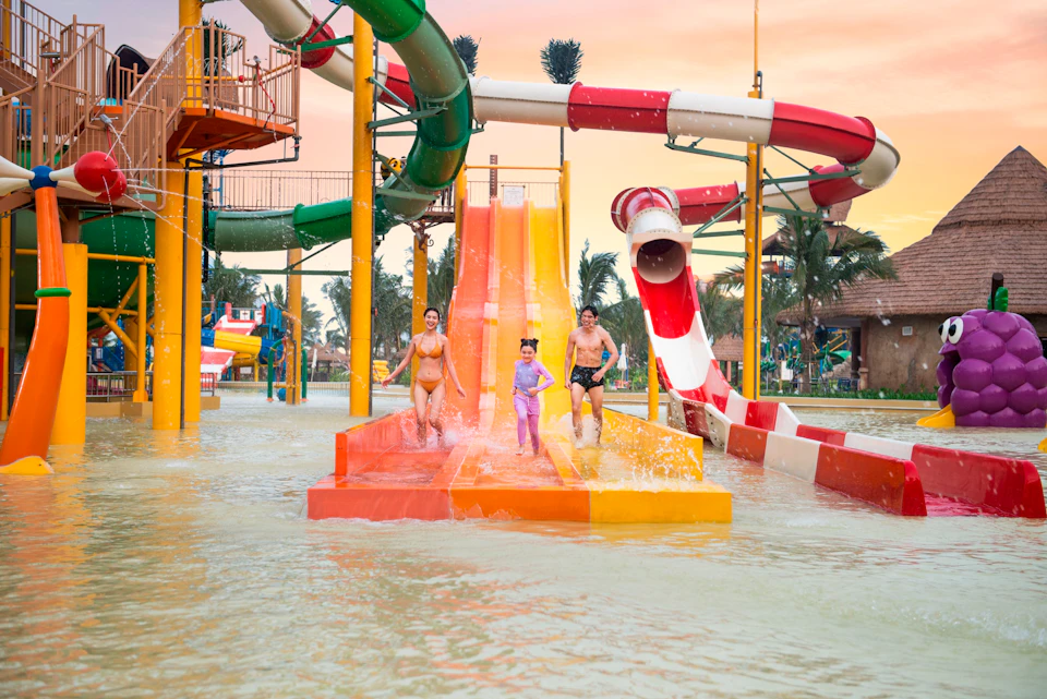 Three people coming down colorful water slides at an outdoor water park with palm trees and a sunset sky in the background.