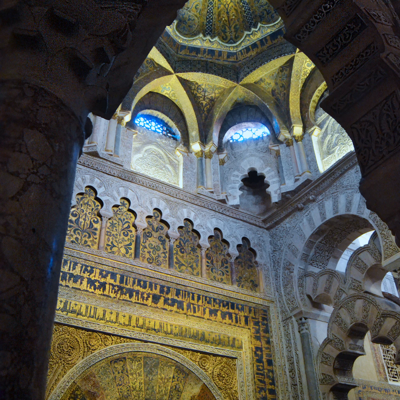 Ornate Islamic architecture with intricate wall carvings, gold accents, and a domed ceiling with small arched windows.