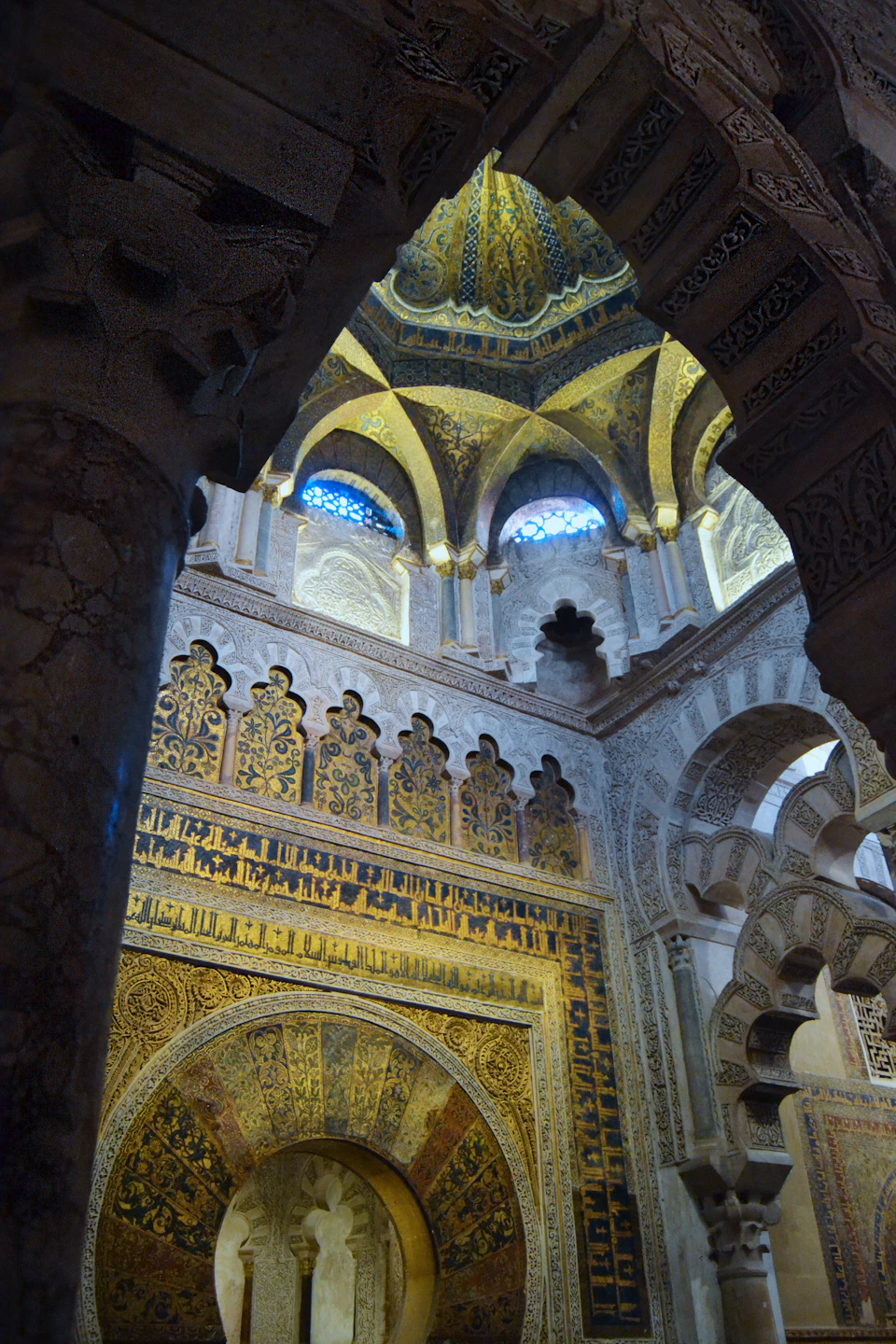 Ornate Islamic architecture with intricate wall carvings, gold accents, and a domed ceiling with small arched windows.