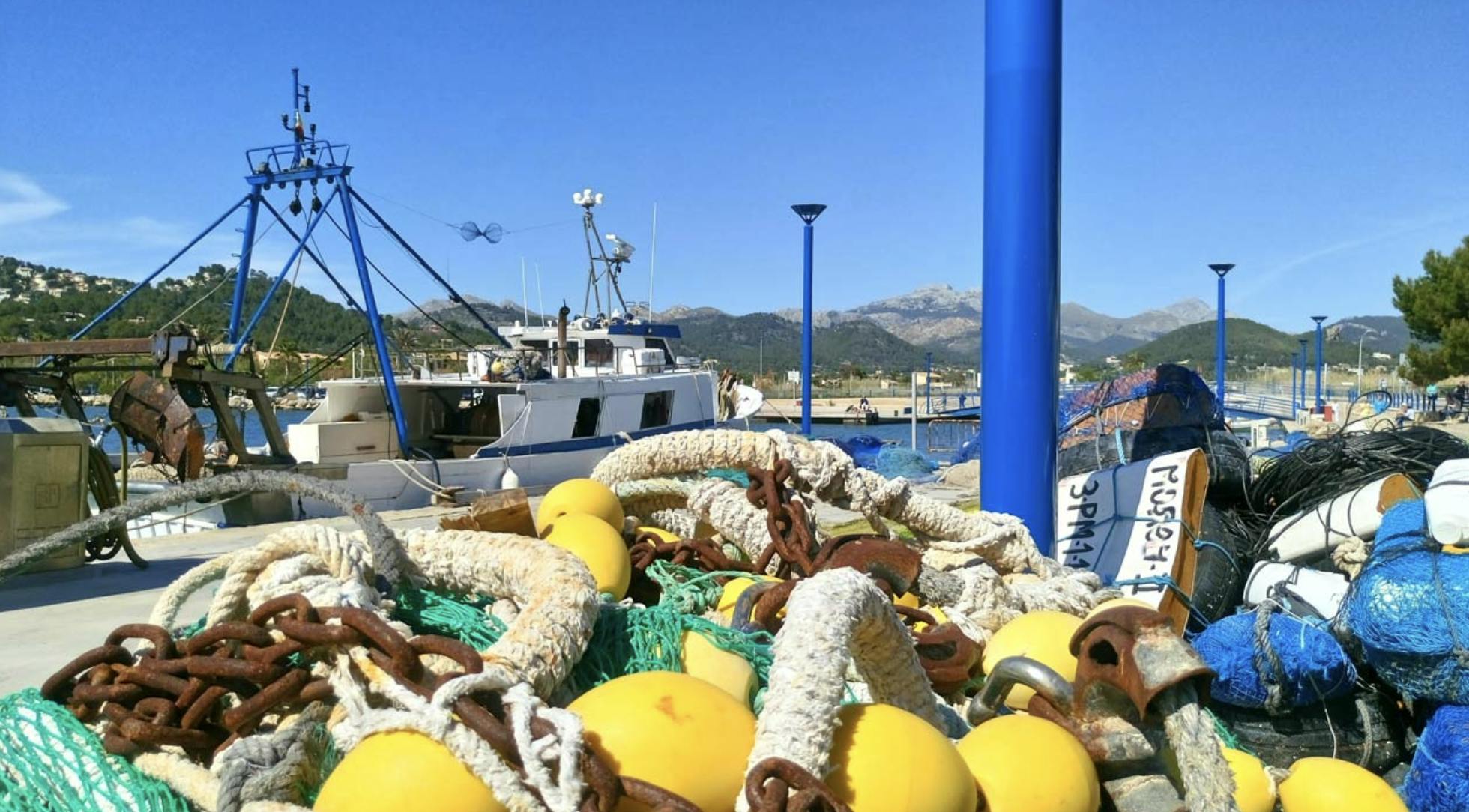 Fischernetze mit gelben Schwimmern auf einem Steg, mit Booten und Bergen im Hintergrund unter einem klaren blauen Himmel.