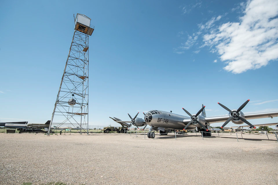 A silver propeller airplane labeled "BF-749" is parked on a gravel surface beside a tall, metal observation tower. Blue sky overhead.