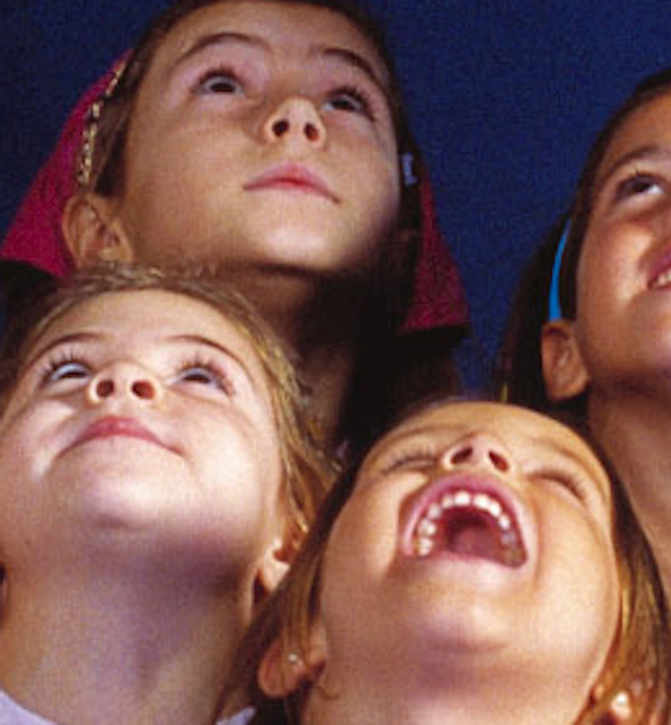 Six children stand inside a large model of a shark's open jaws, looking upward with a dark blue background behind them.