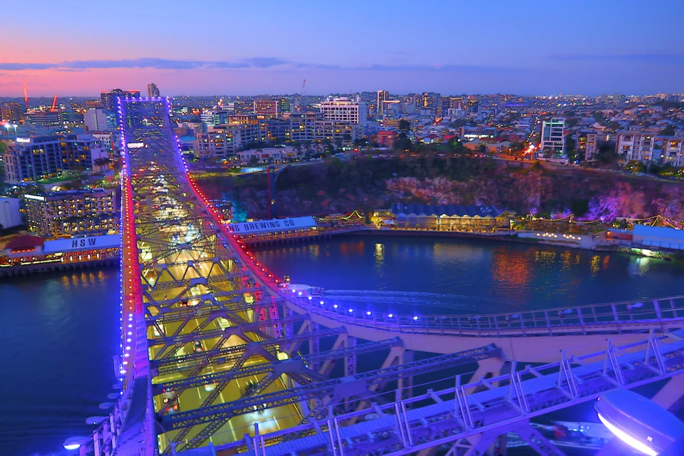 A cityscape at dusk with a lit-up bridge and waterfront area, buildings in the background, and a large body of water.