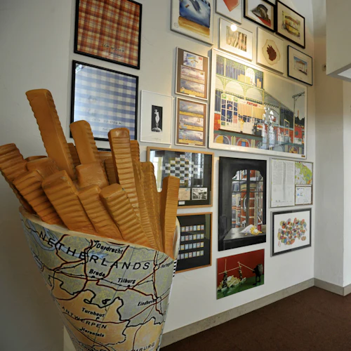 En vägg prydd med olika inramade konstverk. En skulptur som liknar pommes frites i en kartbelagd behållare är placerad framför.