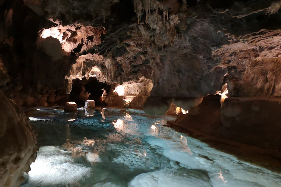 A serene underground cave scene featuring illuminated stalactites and stalagmites reflected in calm, clear water.