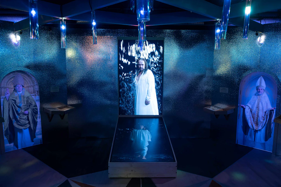 A display featuring a person in a white robe with their reflection, set in a shiny, metallic room with blue lights.
