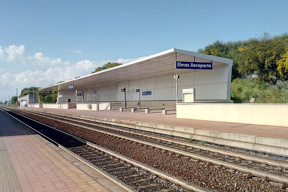 Outdoor train platform labeled "Elmas Aeroporto," with benches, signs, and empty tracks under a clear sky.