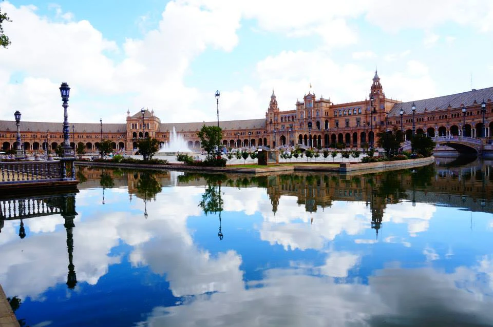 Large historic building with towers and arches, reflected in a calm water body, under a partly cloudy blue sky.