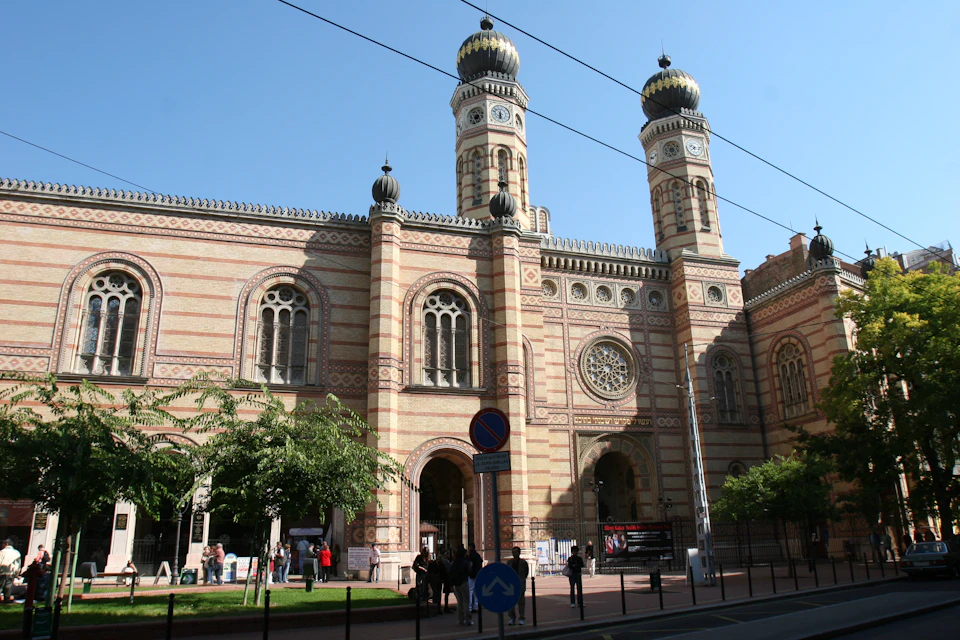 Large brick building with ornate design, two tall domes, arched windows, trees, and a few pedestrians in front under a clear sky.