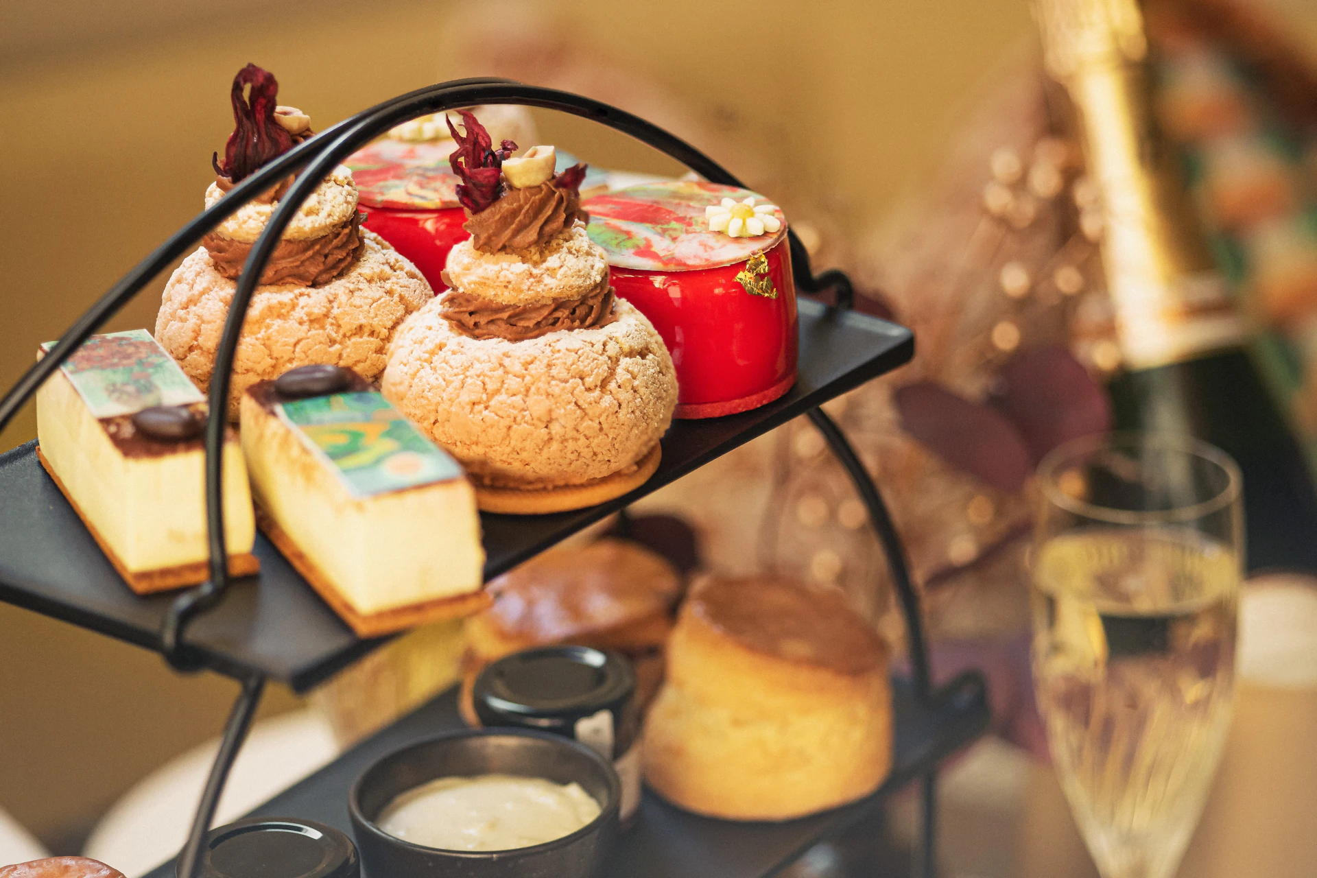 A multi-tiered tray with assorted pastries, including cream puffs, red cakes, scones, and small jars of cream.