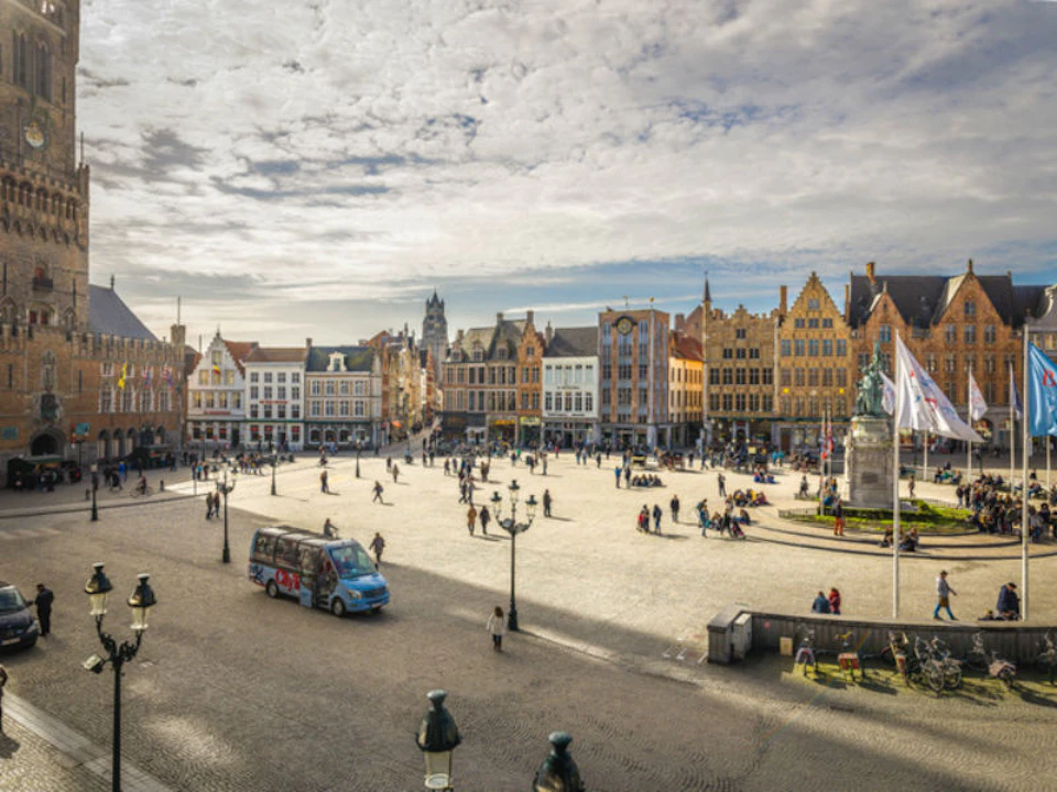 A town square with people walking, a tour bus, a statue with flags, and rows of colorful historic buildings under a partly cloudy sky.