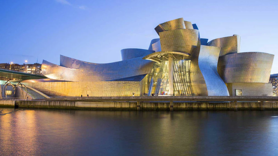 Curved, metallic modern building beside a river at dusk, reflecting golden light. Few people visible near the base.