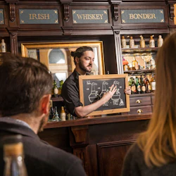 En man förklarar en whiskytillverkningsprocess med hjälp av en tavla i en bar, med två personer som lyssnar i förgrunden.