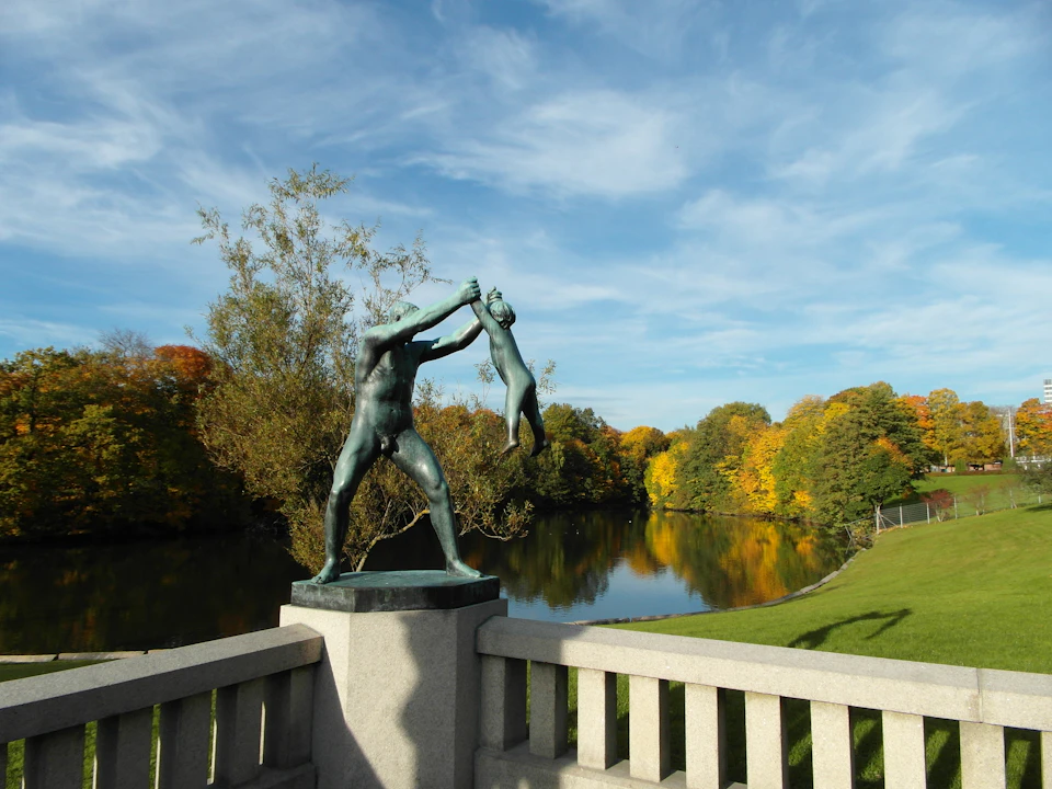 Statue of a man holding a child by its feet, situated on a bridge with a scenic view of a river and colorful autumn trees.