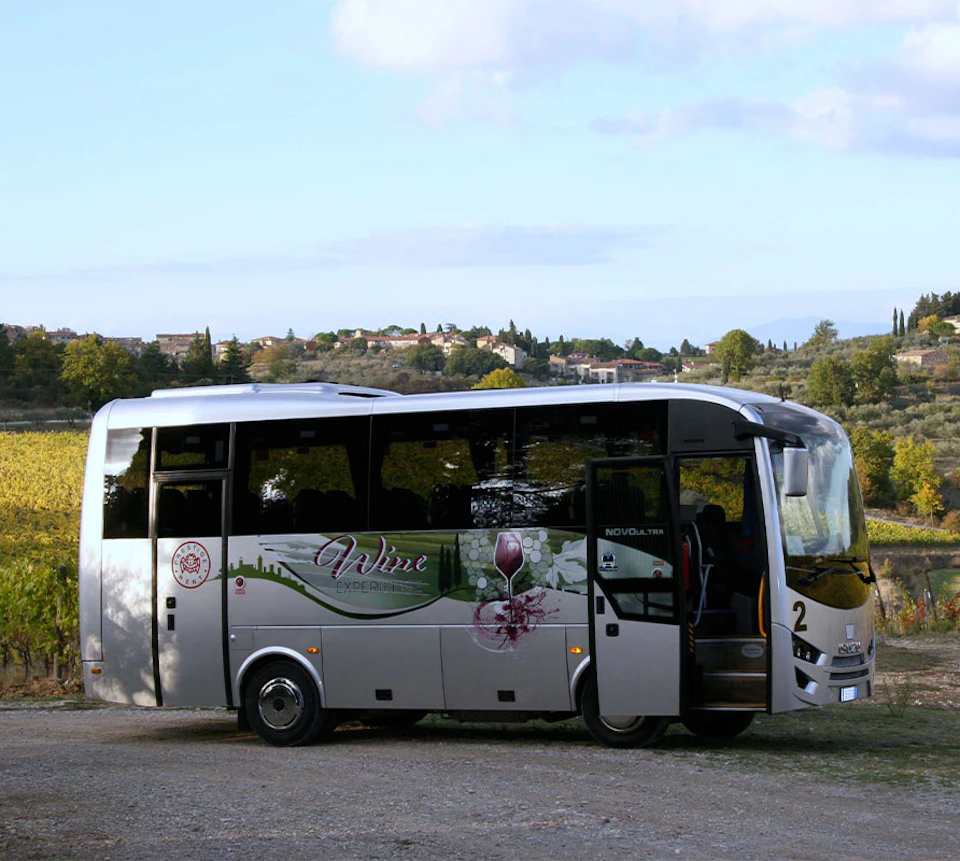 A tour bus with "Wine" branding parked in a scenic countryside with vineyards and a distant village.
