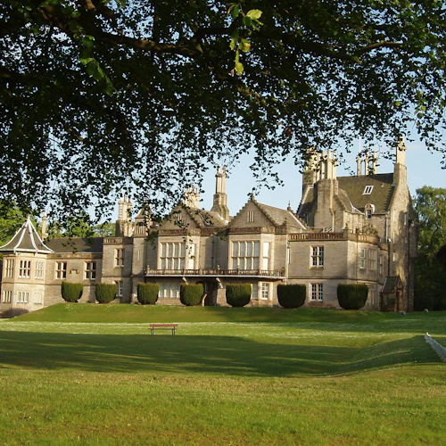 En stor historisk herrgård med välskötta buskar ligger på en frodig grön gräsmatta, inramad av trädgrenar ovanför.