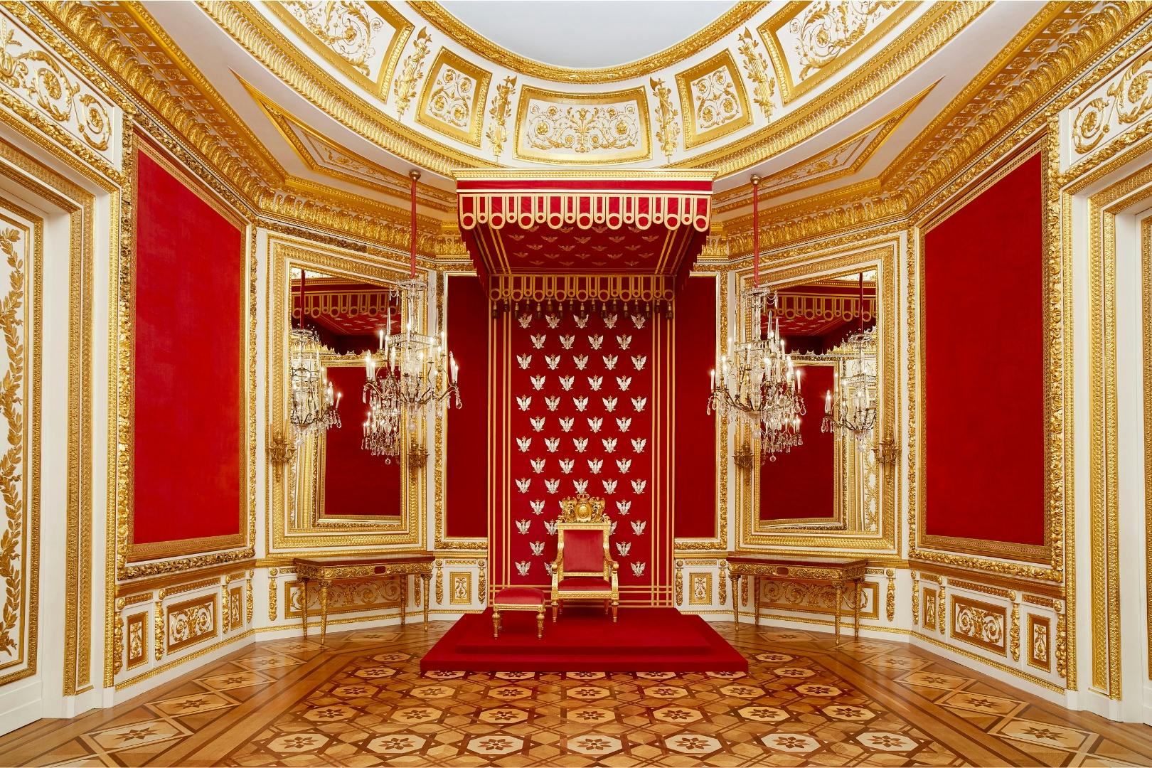 Ein reich verzierter Thronsaal mit rot und gold verzierten Wänden, einem vergoldeten Thron, Kronleuchtern und detaillierten Holzböden.