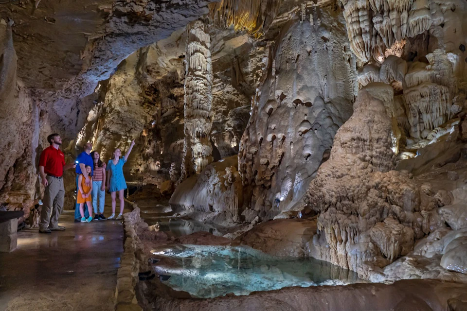 A group of people stands inside a cave with stalactites, stalagmites, and a small illuminated pool on the cave floor.