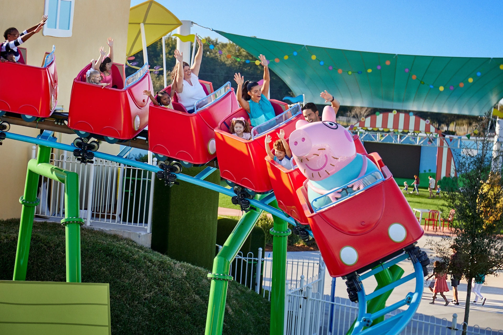 Children and adults riding a Peppa Pig-themed roller coaster with hands raised, smiles, and a colorful canopy in the background.