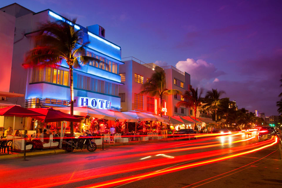 Street with neon-lit hotels, outdoor diners, palm trees, and light trails from passing cars at dusk.