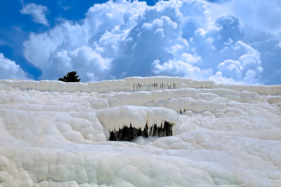 White travertine terraces cascade downwards under a sky filled with fluffy clouds. A small tree is visible at the top left.