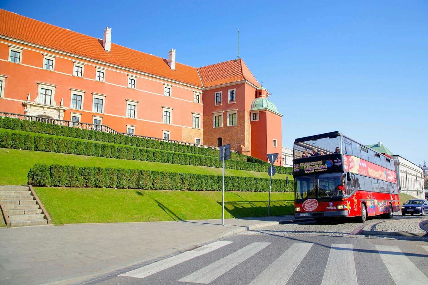 An einem sonnigen Tag fährt ein doppelstöckiger Tour-Bus an einem historischen roten Gebäude mit grünen Hecken davor vorbei.