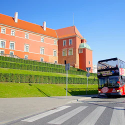 En dubbeldäckad turnébuss passerar en historisk röd byggnad med gröna häckar framför en solig dag.