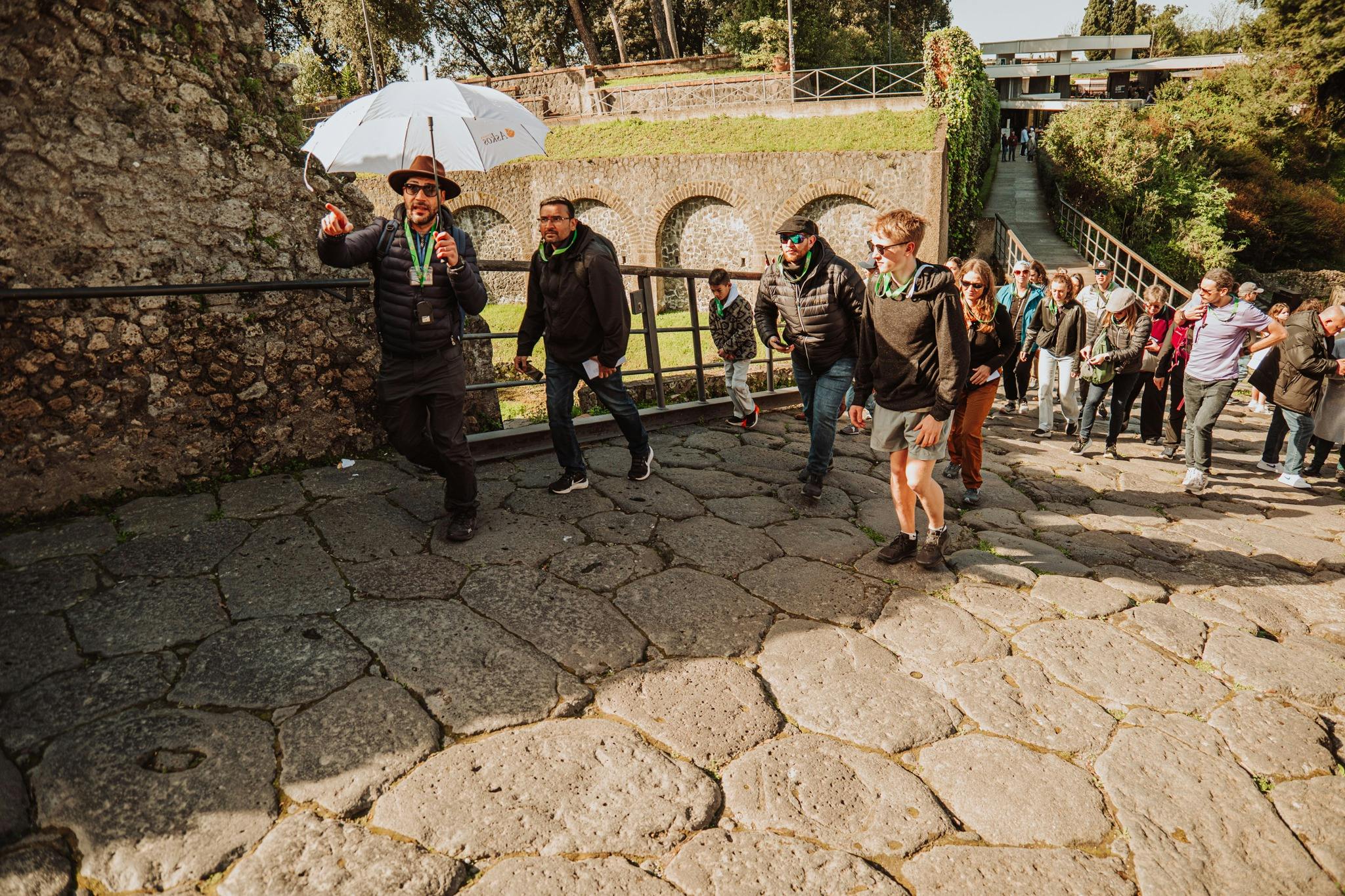 Um guia turístico com um guarda-chuva conduz um grupo de turistas ao longo de um caminho de calçada perto de antigos arcos de pedra à luz do sol.