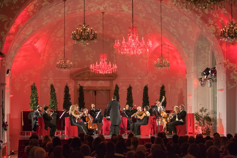 A chamber orchestra performs in an ornately decorated hall with red lighting, chandeliers, and a seated audience.