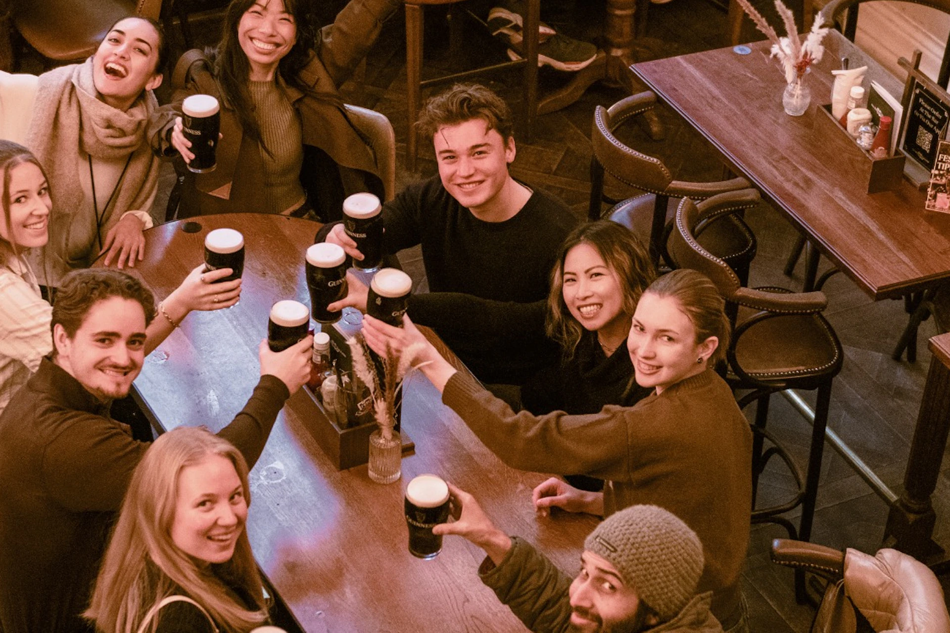 A group of people sitting around a wooden table in a cozy setting, holding up drinks and smiling cheerfully.